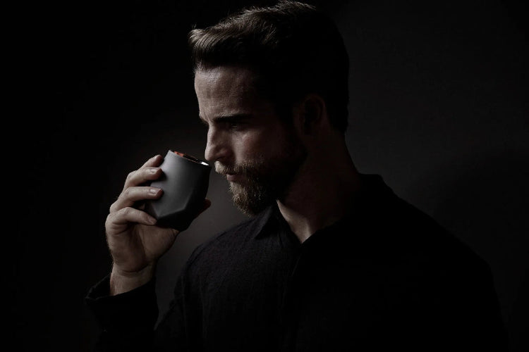 A image of a handsome bearded man against a dark background with a light highlighting his striking features. He hs wearing a black shirt that blends into the background and is holding the Norlan Whisky Glass, VAILD (Black) edition up to his nose to experience the nuanced aromas in his whiskey.
